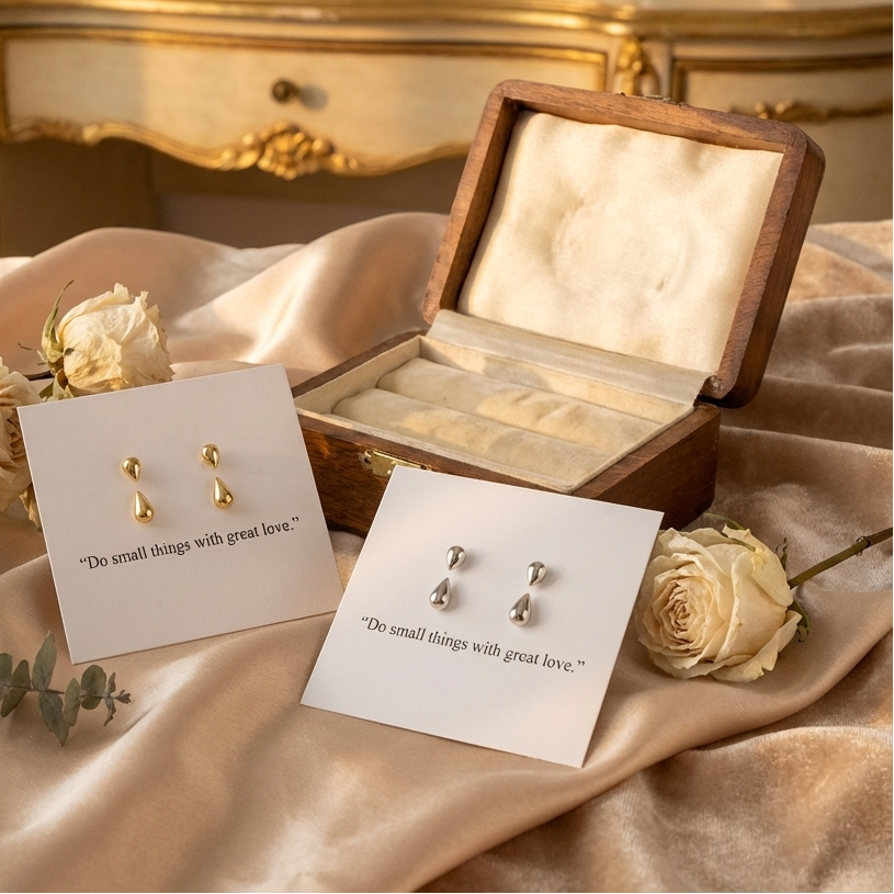 Pair of gold earrings on a card with text, placed on a beige fabric with roses and an ornate wooden box in the background.
